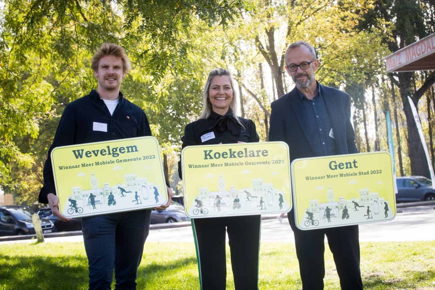 3 mensen poseren met een bord waarop de naam van hun gemeente of stad (Wevelgem, Koekelare, Gent) staat, samen met de tekst 'winnaar meer mobiele gemeente 2022'