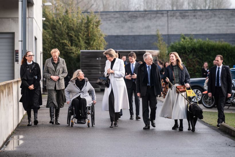 Verschillende mensen lopen in een lijn op een verharde weg, ze zijn met elkaar in gesprek. Onder hen is koningin Mathilde, zij spreekt met een dame in een rolstoel, er is ook een dame met een assistentiehond.