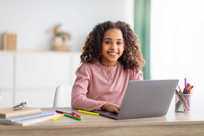 Glimlachend meisje werkt op laptop aan bureau waarop schoolspullen liggen