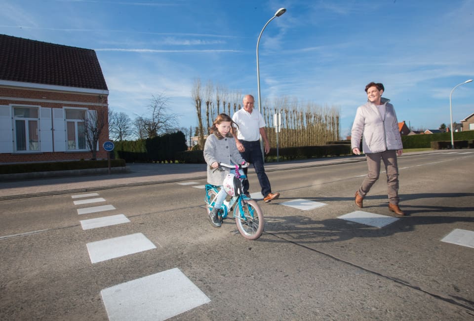 Een meisje steekt de weg over op de fiets. Haar twee grootouders wandelen naast haar.